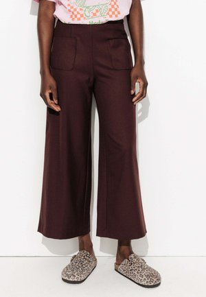 Personne portant un pantalon large marron foncé avec des poches avant et des chaussures à enfiler imprimé léopard, se tenant devant un fond blanc.