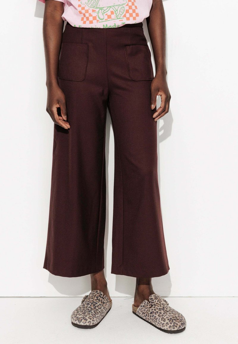 Personne portant un pantalon large marron foncé avec des poches avant et des chaussures à enfiler imprimé léopard, se tenant devant un fond blanc.