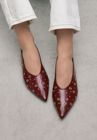 Rode lakleren ballerina's met een spitse neus, voorzien van zilveren oogjes en een gladde, glanzende afwerking. Draagt met witte cropped pants.