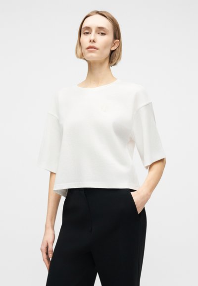 Γυναίκα με κοντά ξανθά μαλλιά, φορώντας άνετο λευκό κοντομάνικο μπλουζάκι και μαύρο παντελόνι, στέκεται με το ένα χέρι στην τσέπη, σε λευκό φόντο.