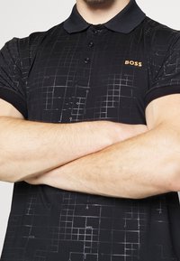 Black polo shirt with a geometric pattern in gray, featuring a three-button placket, a black collar, and a small orange "BOSS" logo.