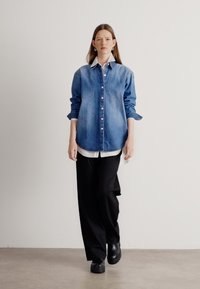 Camicia in denim blu con bottoni, indossata sopra una camicia bianca con colletto, abbinata a pantaloni neri a gamba larga e scarpe nere. Texture lisce.