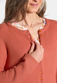 Frau trägt eine strukturierte korallenfarbene Strickjacke mit einer floral gemusterten Bluse darunter und schließt den obersten Knopf. Sichtbares schulterlanges braunes Haar.