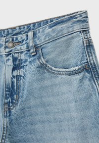 Jean en denim bleu clair en gros plan montrant la poche avant, la boucle de ceinture et le bouton métallique avec des détails de couture visibles.