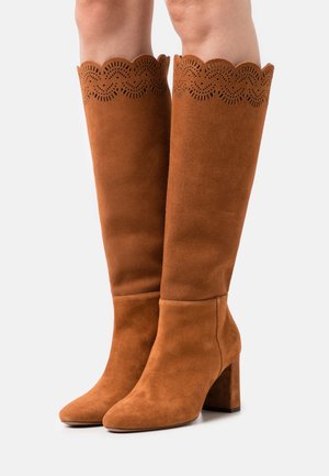 Hnědé semišové kozačky ke kolenům se zvlněným perforovaným horním okrajem, zaoblenou špičkou a čtveratým blokovým podpatkem. Hladký povrch po celém obvodu.
