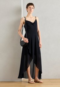 Abito nero senza maniche con scollatura a cappuccio, orlo asimmetrico e sovrapposizione trasparente. La modella tiene una piccola borsa rotonda nera.