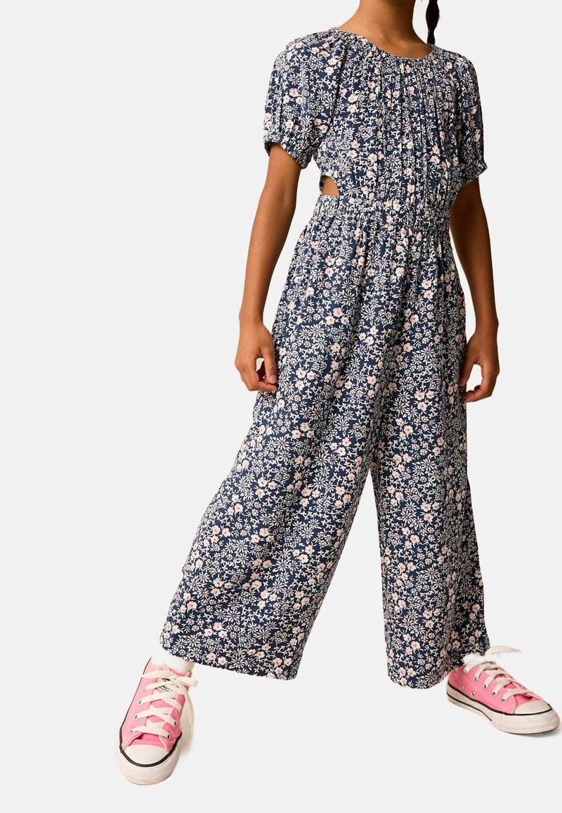 Enfant portant une combinaison marine à motifs floraux avec manches bouffantes et découpes sur les côtés, associée à des baskets basses roses à lacets blancs.