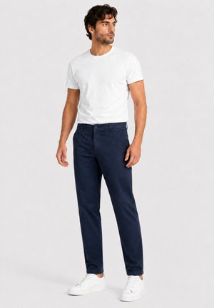 Man draagt een wit t-shirt, donkerblauwe broek en witte sneakers, staat en kijkt naar de zijkant tegen een effen achtergrond.