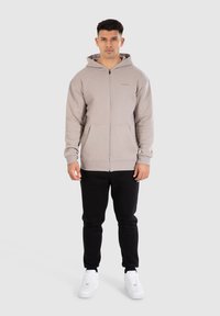 Lichtbeige zip-up hoodie met kangoeroezakken, textuurstof en een capuchon met trekkoord. Gecombineerd met zwarte joggingbroek en witte sneakers.