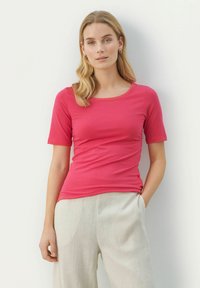 T-shirt aderente a maniche corte in tessuto rosa vivace con scollatura rotonda, abbinato a pantaloni a gamba larga beige chiaro e uno sfondo neutro.