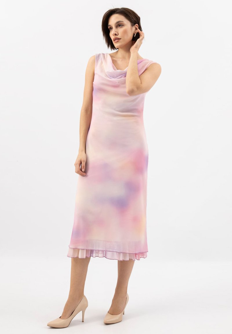 Kobieta w bezrękawnikowej, pastelowej sukience midi z efektem tie-dye, z drapowanym dekoltem, przezroczystym dołem i beżowymi szpilkami, stojąca na tle białego tła.