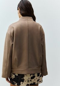 Jachetă din suede artificial maro, cu o croială lejeră, guler, umeri lăsați și o textură netedă, asortată cu o fustă bej cu pete.
