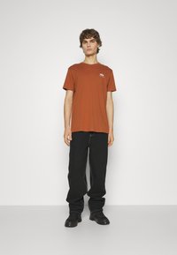 Brun bomulls t-skjorte med korte ermer og rund hals, kombinert med svarte bukser og svarte sko; minimalistisk design.