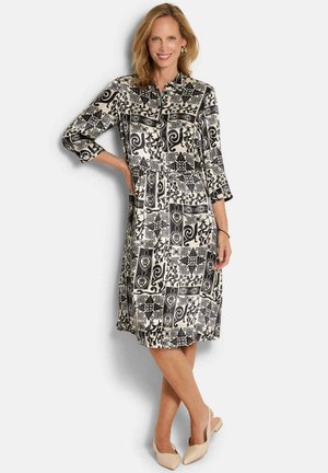 Frau lächelnd, trägt ein knieumspielendes schwarz-weiß gemustertes Kleid mit dreiviertellangen Ärmeln und beigefarbenen spitzen Schuhen, steht mit einer Hand in der Hüfte.