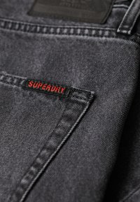 Tissu en denim noir avec une texture subtile, comportant une étiquette rouge "SUPERDRY" sur la poche et une étiquette sombre au-dessus.