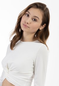 Top bianco a maniche lunghe con dettagli a nodo attorcigliato in vita. Tessuto liscio e design aderente. Linee semplici e pulite.