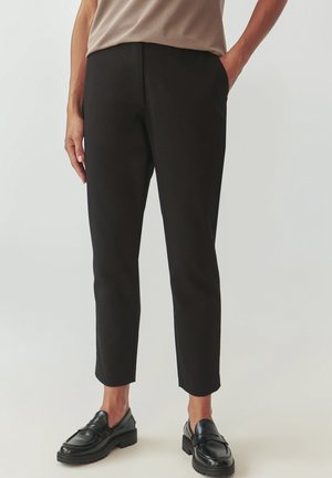 Pantalones negros ajustados con un material suave, diseño slim, longitud recortada y bolsillos laterales, combinados con mocasines negros pulidos.