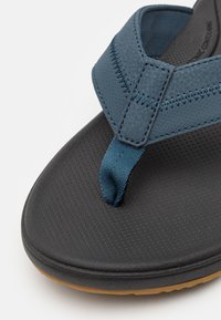 Blå flip-flop sandal med en teksturert svart såle, med en blå stropp med stikking og en liten tærskille.