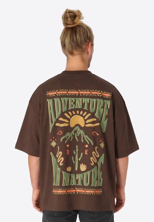 Man met blond haar in een knot, draagt een bruin oversized T-shirt met een kleurrijke "Avontuur in de natuur" afbeelding van zon, bergen, cactus en slangen.