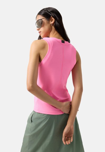 Frau, die große Sonnenbrillen, ein ärmelloses, geripptes rosa Oberteil und einen grünen Rock trägt, von hinten vor hellem, einfarbigem Hintergrund gezeigt.