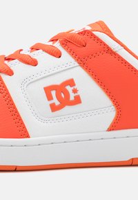 Oranžové a biele tenisky so syntetickou kožou na vrchu, obsahujúce gumovú podrážku, ploché šnúrky a akcent DC loga na boku.