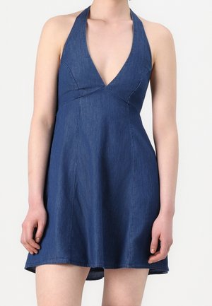 Femme portant une robe en denim bleu sans manches à col halter en V, taille cintrée et jupe évasée.