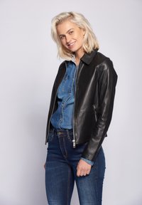 Zwarte leren jas met een kraag, ritsluiting en twee zijzakken, gedragen over een denim shirt, gecombineerd met blauwe spijkerbroeken.
