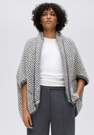Femme aux cheveux bouclés portant une chemise blanche, un pantalon gris taille haute et un châle tricoté gris et blanc à motifs.