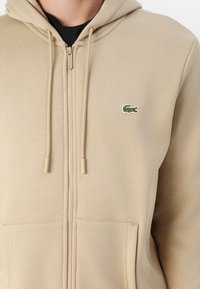 Felpa beige con zip, dotata di tasca a marsupio, tessuto testurizzato, cappuccio con cordino e un piccolo logo di coccodrillo verde sul lato sinistro del petto.