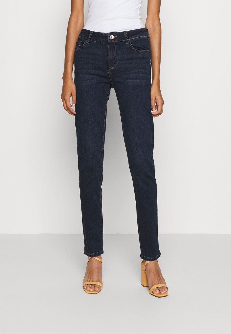 Jeans de mezclilla oscuros de corte slim con un diseño clásico de cinco bolsillos, que incluyen cierre de cremallera, botón y detalles de costura en contraste.