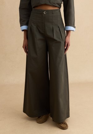 Pantalones de pierna ancha de talle alto en color verde oliva con pliegues, combinados con una chaqueta corta que deja al descubierto una pequeña parte del abdomen y zapatos estilo mocasín.