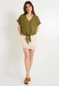 Top de color verde caqui de manga corta con escote en forma de V y detalle de botones, combinado con una falda mini de encaje beige con dobladillo en forma de ola. Zapatos marrones de slip-on.