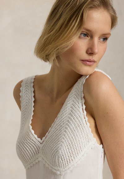 Jauna sieviete ar īsiem blondiem matiem, valkā baltu bezroku tamborētu topu, skatās sānis ar neitrālu izteiksmi.