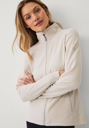 Vrouw met lang blond haar, gekleed in een beige fleecejack met een ritssluiting aan de voorkant, staand met gekruiste armen tegen een neutrale achtergrond.