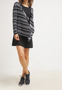 Černé šaty doplněné vzorovaným černobílým kardiganem. Kardigan má dlouhé rukávy a otevřený střih. Celkový vzhled doplňují černé tenisky.