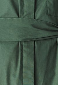 Tume roheline kangas sileda tekstuuriga, millel on voldid ja kõhule seotud kangast vöö. Pinna õmblused on nähtavad õmbluste kõrval.