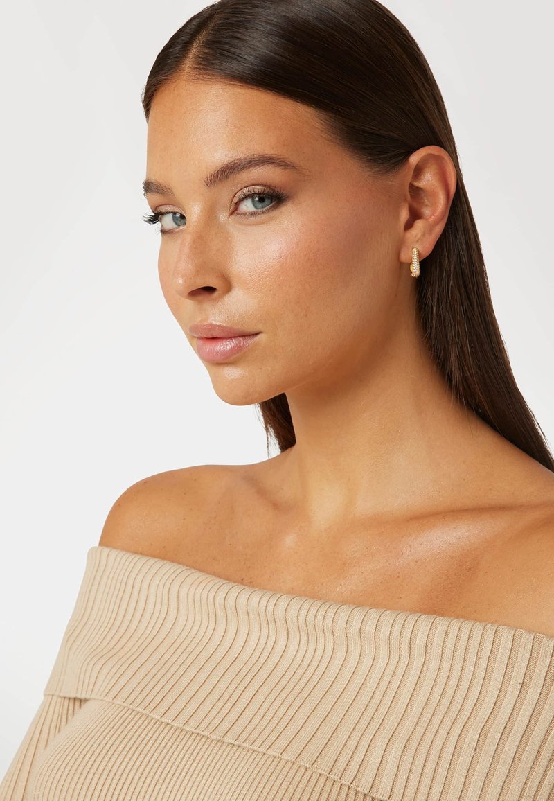 Boucles d'oreilles en or avec une surface texturée, portées avec un haut beige côtelé à épaules dénudées, mettant en avant une peau douce et lisse ainsi que de longs cheveux.