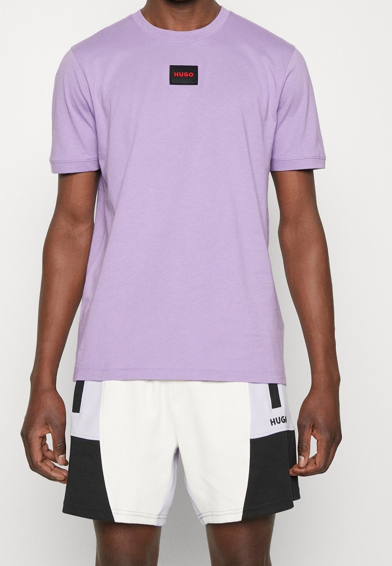 Homme portant une chemise à manches courtes lavande avec un patch logo rouge "HUGO" et un short bicolore blanc, noir et lavande avec le texte "HUGO".