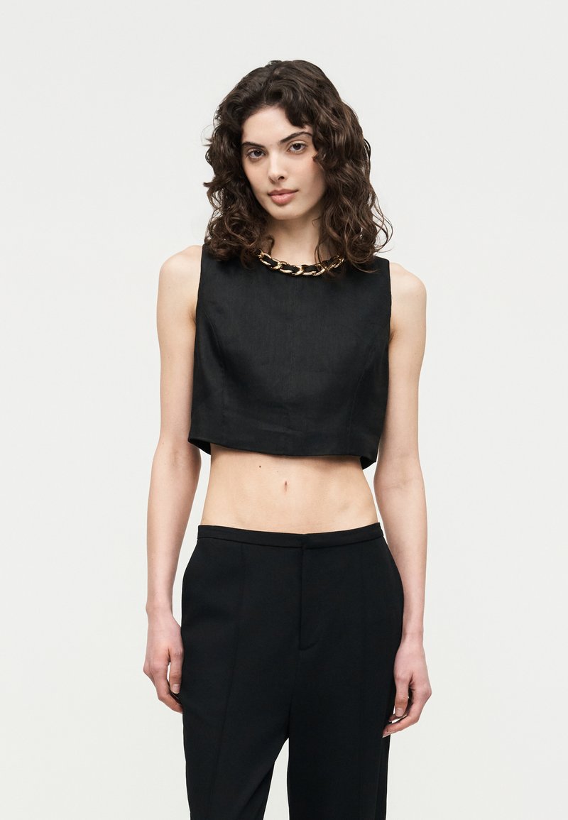 Γυναίκα με σγουρά μαλλιά που φοράει μαύρο κοντό τοπ χωρίς μανίκια, με χρυσή αλυσίδα στο λαιμό και μαύρο ψηλόμεσο παντελόνι, στέκεται μπροστά σε λευκό φόντο.