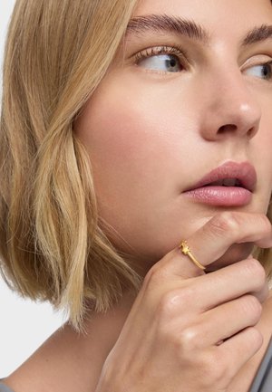 Donna con capelli biondi corti che guarda di lato, appoggiando il mento sulla mano, indossando un anello d'oro con un motivo a stella.