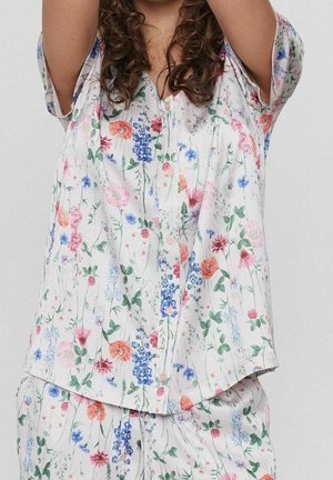 Femme portant un ensemble de pyjama blanc à imprimé floral multicolore, les bras levés, cheveux bruns bouclés partiellement visibles sur un fond uni.