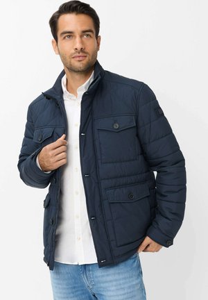 Uomo che indossa una giacca trapuntata blu scuro sopra una camicia bianca e jeans blu, che sistema la cerniera della giacca, guardando leggermente verso sinistra su sfondo bianco.