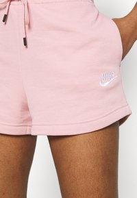 Růžové bavlněné šortky s elastickým pasem a šňůrkou, na kterých je bílý logo Nike. Hladký povrch, obroubené okraje a viditelné boční kapsy.