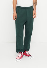 Green cotton jogger pants with an elastic waistband, featuring a relaxed fit and side pockets, paired with red sneakers.