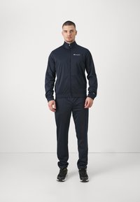 Tuta navy in tessuto morbido. La giacca presenta una chiusura con zip, colletto alto e logo sul petto. I pantaloni hanno polsini elastici.