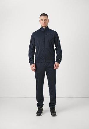 Tuta navy in tessuto morbido. La giacca presenta una chiusura con zip, colletto alto e logo sul petto. I pantaloni hanno polsini elastici.