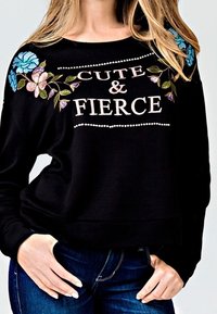 Sweatshirt noir court avec motif de fleurs brodées en bleu et rose sur les épaules, et le texte "CUTE & FIERCE" mis en avant en blanc et rose.