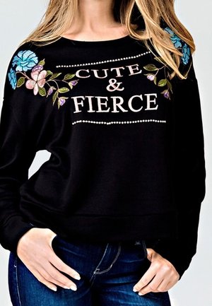 Felpa nera corta con fiori ricamati blu e rosa sulle spalle, con la scritta "CUTE & FIERCE" evidenziata in bianco e rosa.