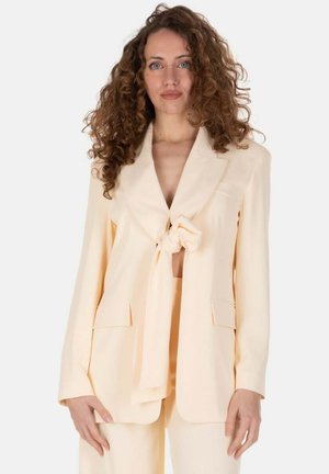 Donna con capelli ricci che indossa una giacca da completo color crema annodata davanti sopra pantaloni abbinati, in piedi contro uno sfondo semplice.