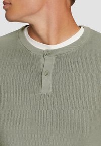 Lichtgroen gestructureerd henley-shirt met een ronde halslijn, voorzien van twee knopen en een wit shirt eronder.